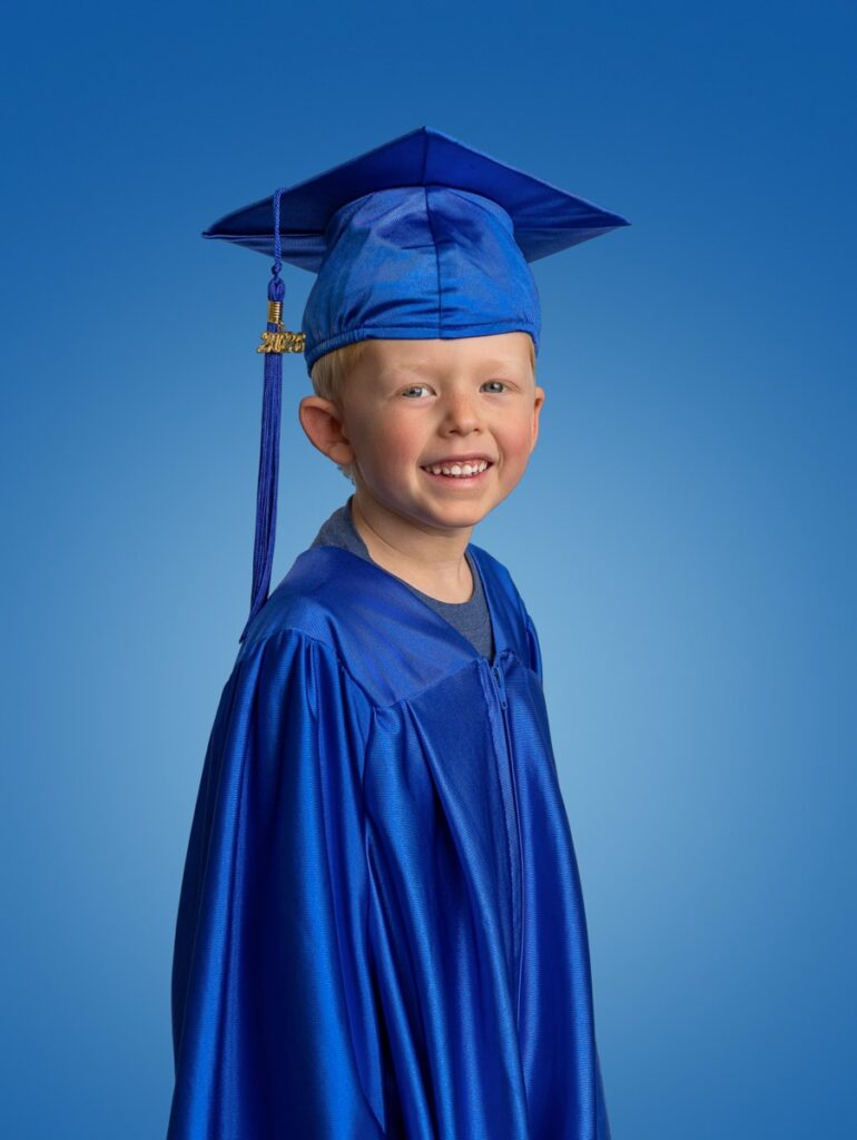 Cap and gown preschool graduation portrait of a boy wearing a blue robe  cap and with a gold tassel smiling for the camera. 